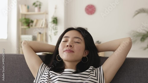 Young asian woman with hands behind head relaxing on sofa at home. Joyful chinese ethnicity female daydreaming on couch