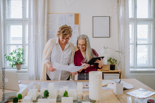 Mature women eco architects with model of modern bulidings and blueprints working together in office.