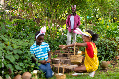 Happy african american siblings in bunny ears hiding easter eggs with grandfather in backyard