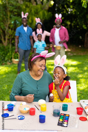 Happy african american girl and grandmother painting easter eggs while family in background