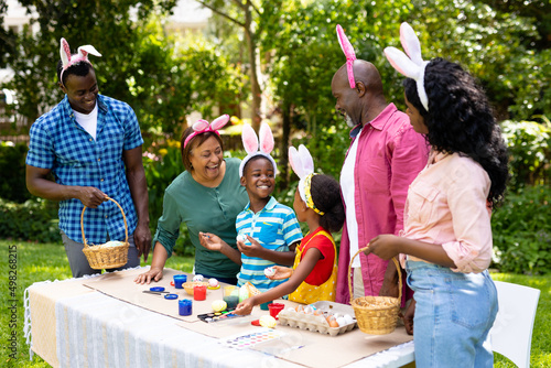 Happy african american multigenerational family wearing bunny ears painting easter eggs in backyard