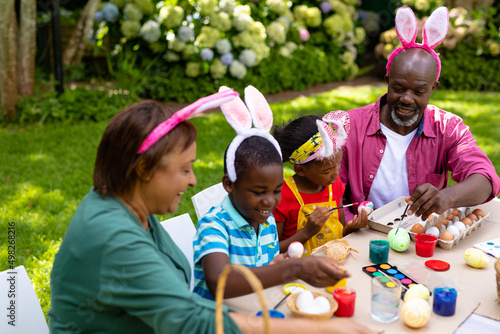Happy african american siblings and grandparents in bunny ears painting eggs on easter day