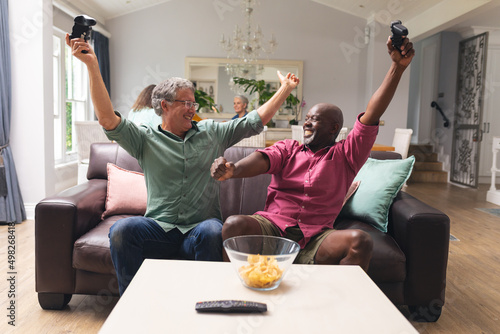 Multiracial senior male friends with arms raised enjoying video game at home