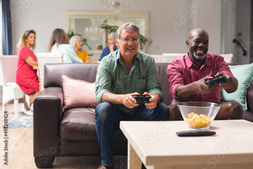 Multiracial senior male friends playing video game while females in background at home