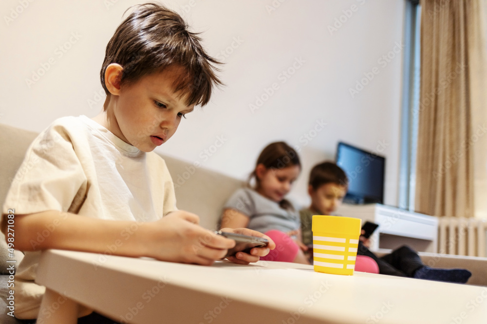 Three kids are sitting on couch and playing with modern mobile devices. Small group of children sit on sofa and use technology at home during the day. Group of kids using wireless technology at home. Three kids are sitting on couch and playing with modern mobile devices. Small group of children sit on sofa and use technology at home during the day. Group of kids using wireless technology at home.
