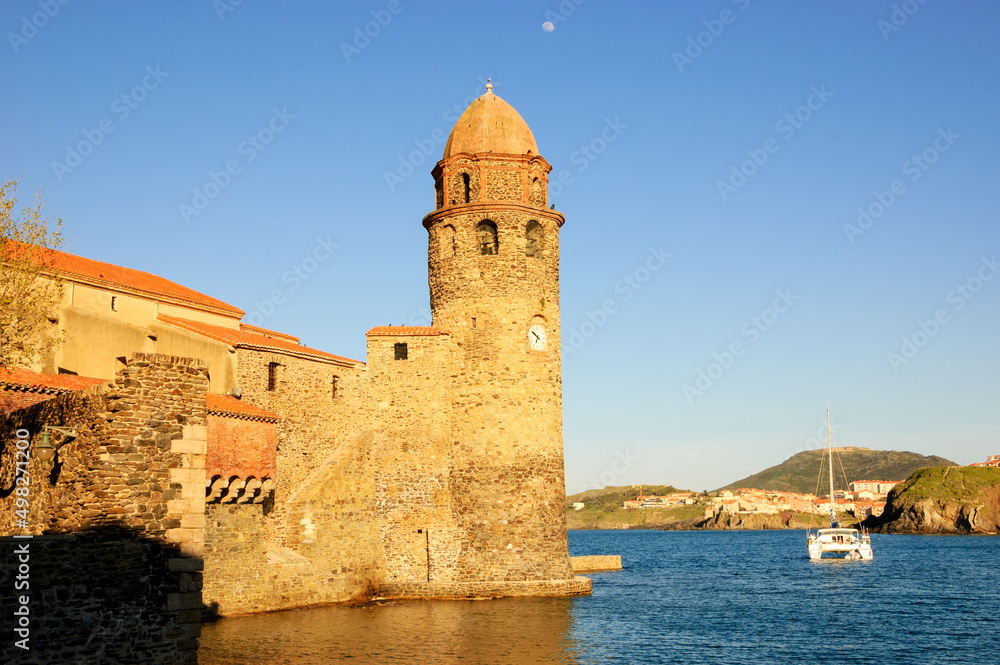 The Church of Our Lady of the Angels in the port of Collioure, France. Moon in the sky and white sailing boat. Sunset.