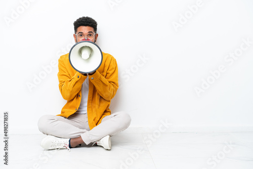 Young Brazilian man sitting on the floor isolated on white background shouting through a megaphone