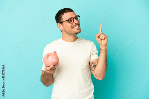 Brazilian man holding a piggybank over isolated blue background pointing up a great idea