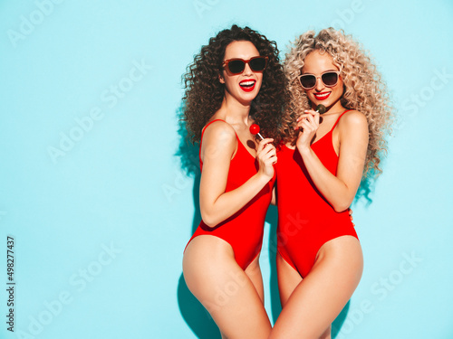 Two beautiful smiling hipster women in red swimwear bathing suits.Trendy models with curls hairstyle in studio. Hot female posing near wall in sunglasses.Holding lollipop. Isolated on blue