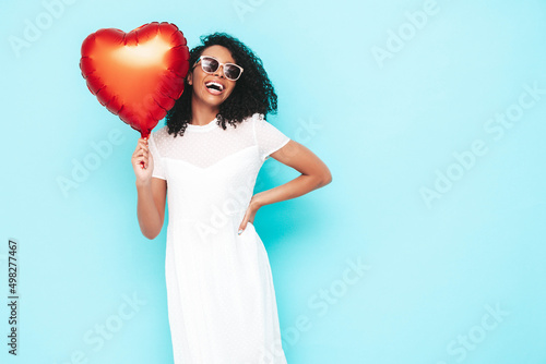 Beautiful black woman with afro curls hairstyle. Smiling model dressed in white summer dress. Sexy carefree female posing near blue wall in studio. Tanned and cheerful. Holding heart air balloon © halayalex
