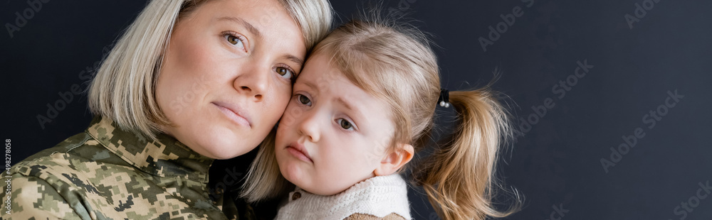 blonde military woman looking at camera near sad daughter isolated on black, banner. blonde military woman looking at camera near sad daughter isolated on black, banner.