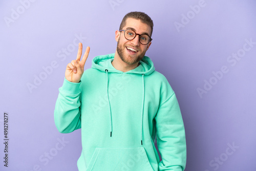 Young handsome caucasian man isolated on purple background smiling and showing victory sign © luismolinero