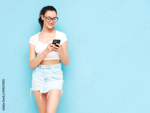 Young beautiful smiling female in trendy summer jeans skirt. Sexy carefree woman posing near blue wall in studio. Positive brunette model looking at cellphone screen. Holding smartphone and using apps