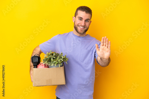 Young caucasian making a move while picking up a box full of things isolated on yellow background saluting with hand with happy expression