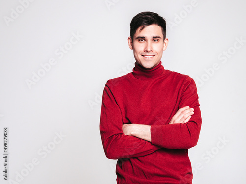 Portrait of handsome smiling model. Sexy stylish man dressed in red turtleneck sweater and jeans. Fashion hipster male posing on grey background in studio. Isolated. Crossed arms