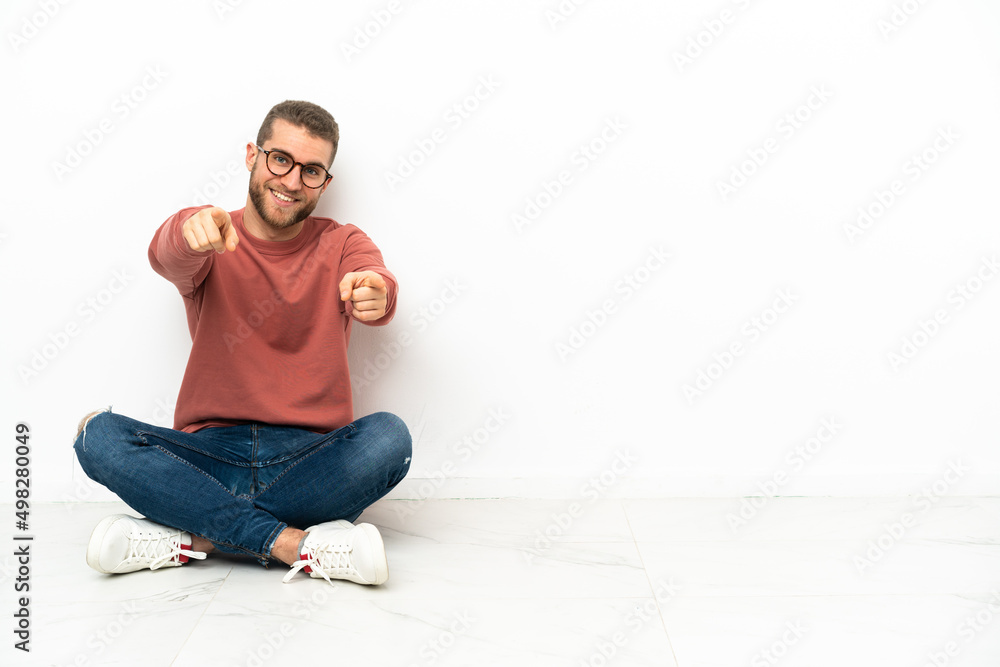 Young handsome man sitting on the floor points finger at you while smiling Young handsome man sitting on the floor points finger at you while smiling