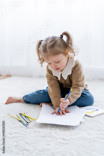 little girl with ponytails drawing on floor near card with blue and yellow heart.