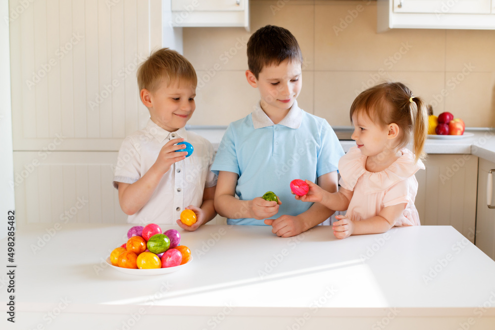 Sibling children in a bright kitchen with colored eggs celebrate Easter.
