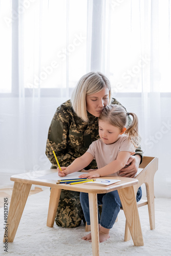 woman in military uniform hugging little daughter drawing with blue and yellow pencils.