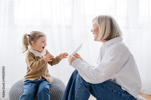 cheerful blonde woman holding card near amazed daughter while sitting on poufs at home. photo