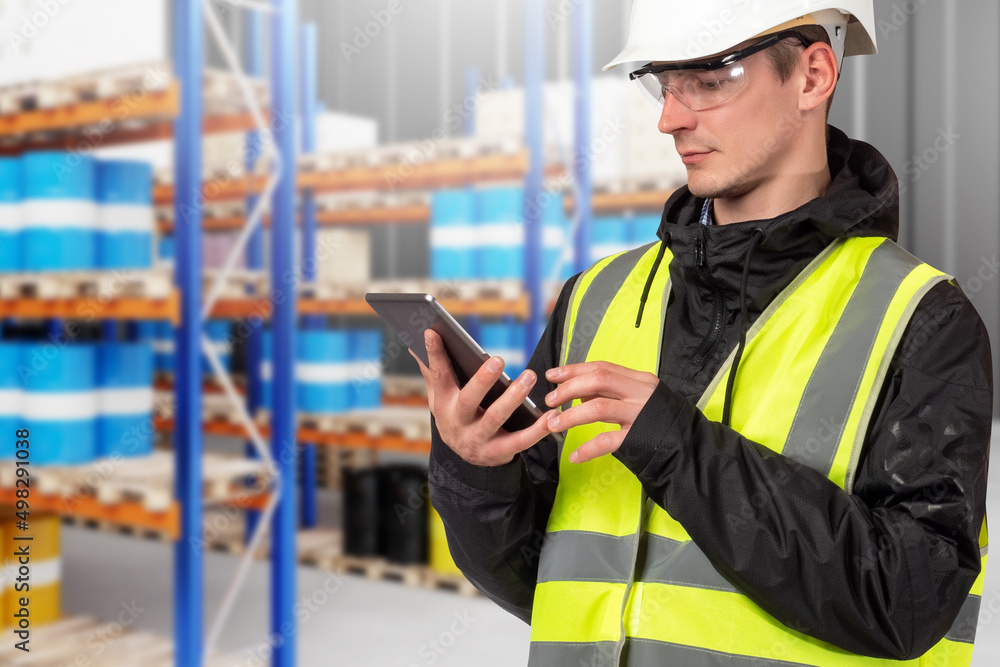 Man in a fuel warehouse. Storekeeper with an electronic tablet in his hands. Blurred shelves with barrels as a background. Storage of liquids in a warehouse. Storage of fuel and chemicals. Man in a fuel warehouse. Storekeeper with an electronic tablet in his hands. Blurred shelves with barrels as a background. Storage of liquids in a warehouse. Storage of fuel and chemicals.