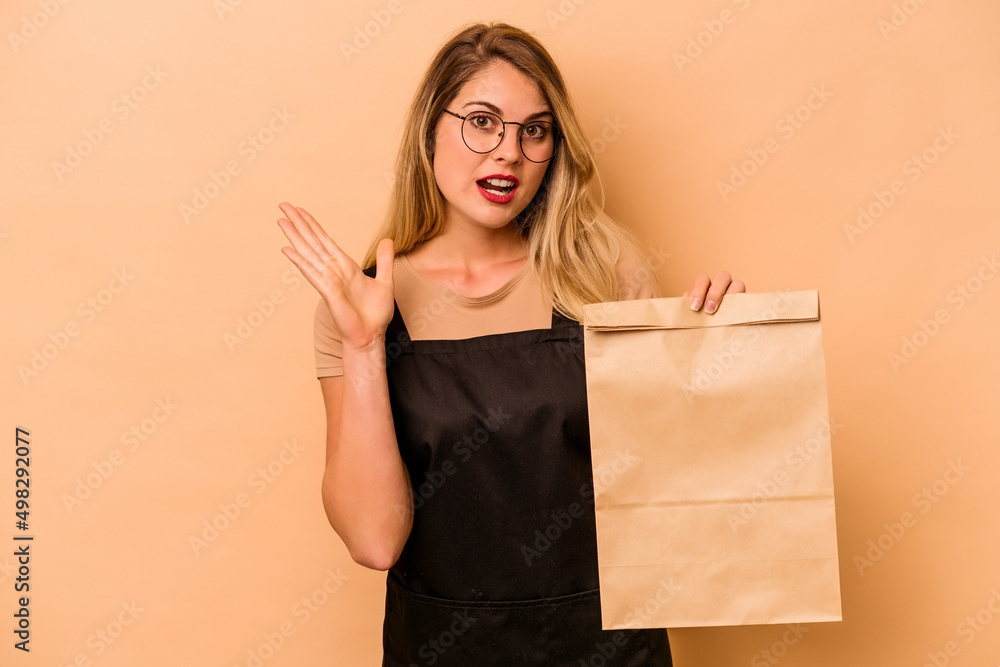 Restaurant waiter caucasian woman holding a take away bag isolated on beige background surprised and shocked. Restaurant waiter caucasian woman holding a take away bag isolated on beige background surprised and shocked.