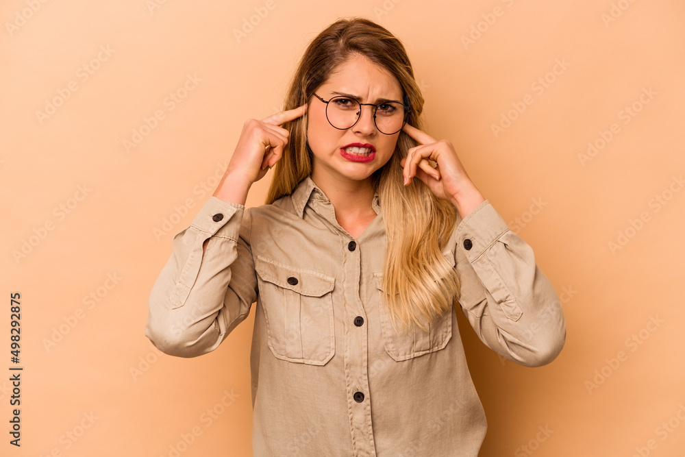 Young caucasian woman isolated on beige background covering ears with fingers, stressed and desperate by a loudly ambient. Young caucasian woman isolated on beige background covering ears with fingers, stressed and desperate by a loudly ambient.