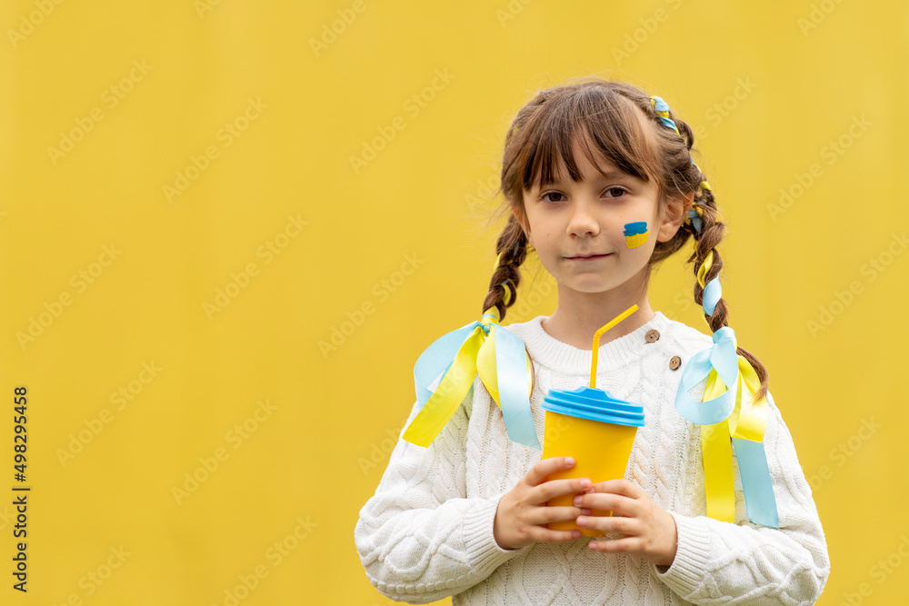 A little Ukrainian girl with two pigtails and yellow and blue ribbons drinks a hot drink from a glass of patriotic colors like the Ukrainian flag. Stop the war in Ukraine. Volunteer Help for Refugees A little Ukrainian girl with two pigtails and yellow and blue ribbons drinks a hot drink from a glass of patriotic colors like the Ukrainian flag. Stop the war in Ukraine. Volunteer Help for Refugees