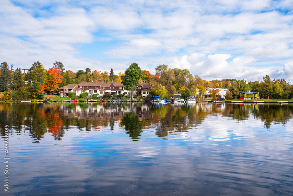 View of lakeside brick row houses surrounded by deciduous trees at the peak of fall foliage on a partly cloudy morning. Reflection in water.
