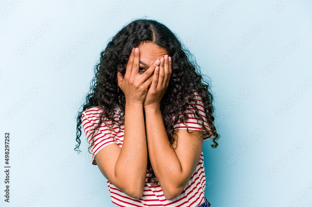 Young hispanic woman isolated on blue background blink at the camera through fingers, embarrassed covering face. Young hispanic woman isolated on blue background blink at the camera through fingers, embarrassed covering face.