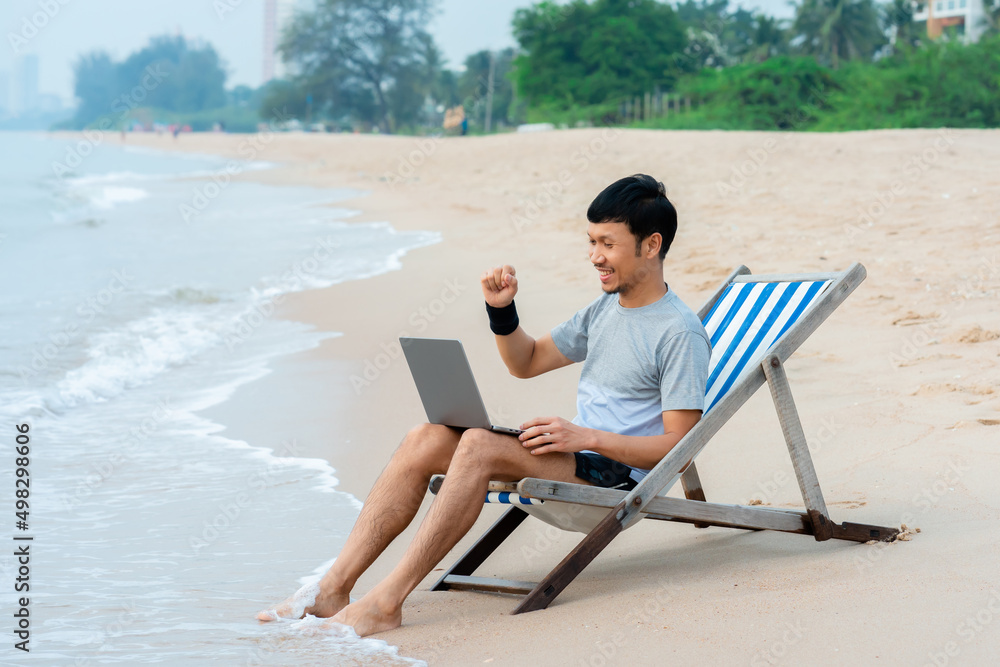 A handsome Asian man sits on a chair in a laptop computer by the sea as the waves crash against the shore, he raises his hand glad to offer the job, pleasantly by the sea, work from home.