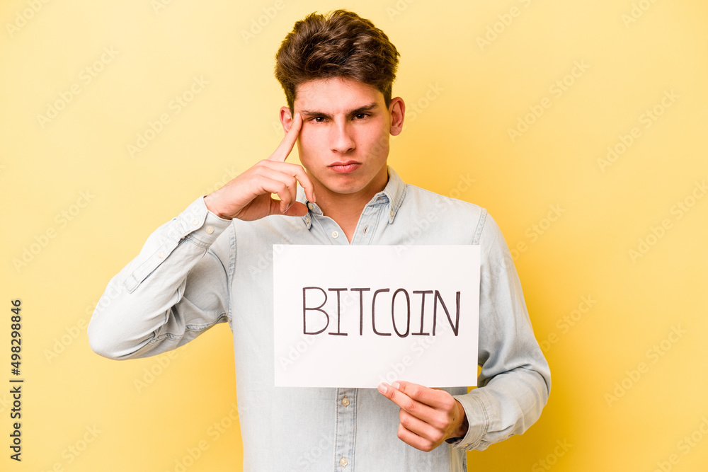 Young caucasian man holding bitcoin placard isolated on yellow background pointing temple with finger, thinking, focused on a task. Young caucasian man holding bitcoin placard isolated on yellow background pointing temple with finger, thinking, focused on a task.