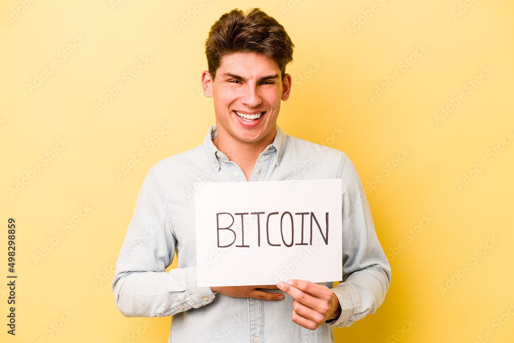 Young caucasian man holding bitcoin placard isolated on yellow background laughs out loudly keeping hand on chest. Young caucasian man holding bitcoin placard isolated on yellow background laughs out loudly keeping hand on chest.
