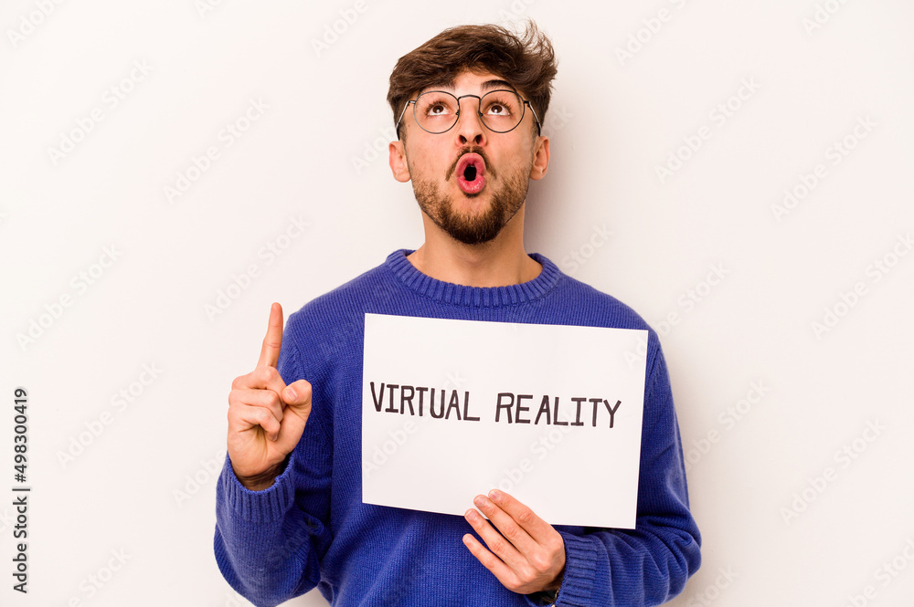 Young hispanic man holding a virtual reality placard isolated on white background pointing upside with opened mouth. Young hispanic man holding a virtual reality placard isolated on white background pointing upside with opened mouth.