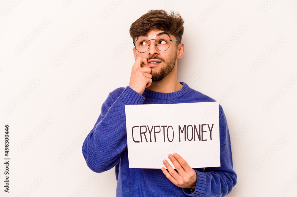 Young hispanic man holding a crypto money placard isolated on white background relaxed thinking about something looking at a copy space. Young hispanic man holding a crypto money placard isolated on white background relaxed thinking about something looking at a copy space.