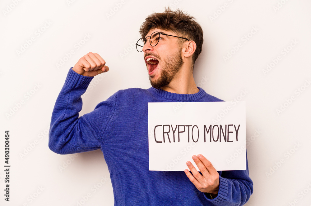 Young hispanic man holding a crypto money placard isolated on white background raising fist after a victory, winner concept. Young hispanic man holding a crypto money placard isolated on white background raising fist after a victory, winner concept.