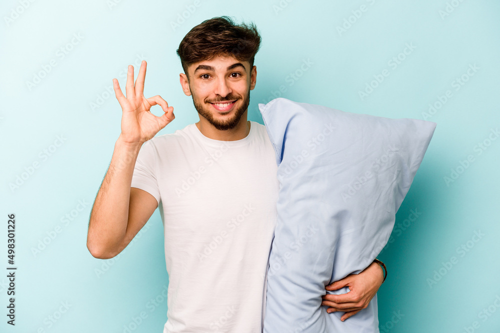 Young hispanic man wearing a pajama holding pillow isolated on blue background cheerful and confident showing ok gesture. Young hispanic man wearing a pajama holding pillow isolated on blue background cheerful and confident showing ok gesture.