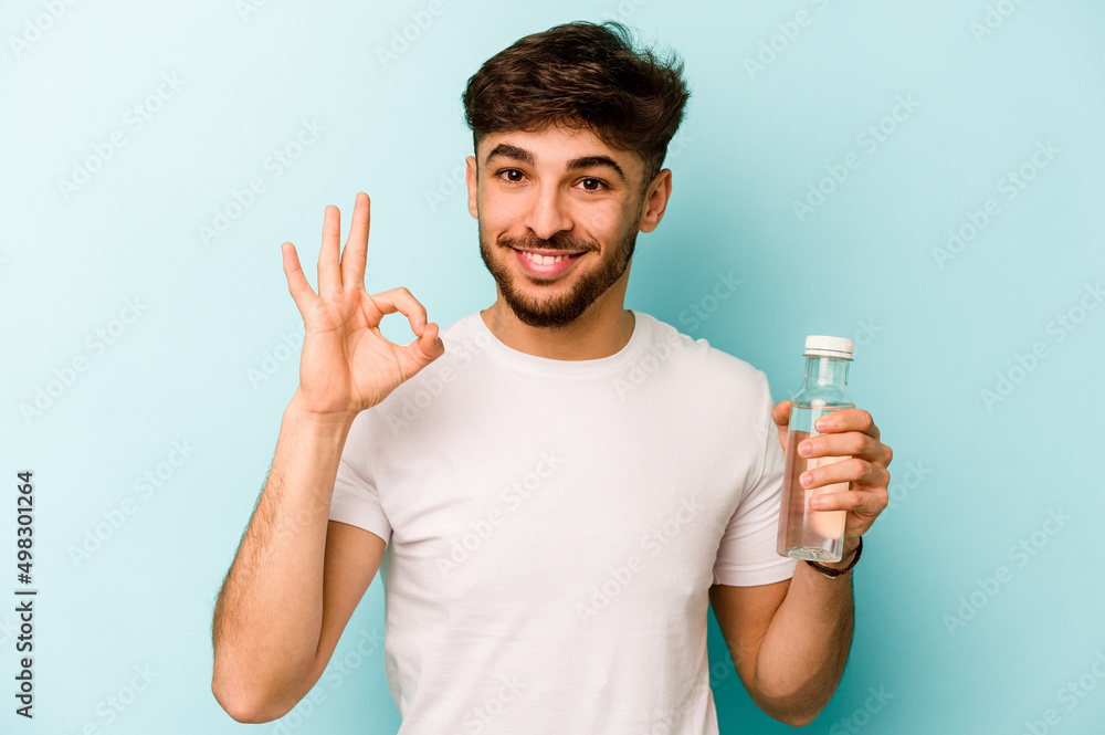 Young hispanic man holding a bottle of water isolated on white background cheerful and confident showing ok gesture. Young hispanic man holding a bottle of water isolated on white background cheerful and confident showing ok gesture.