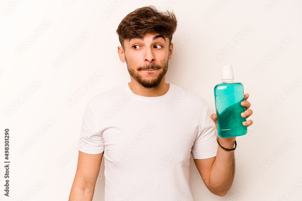 Young hispanic man holding mouthwash isolated on white background confused, feels doubtful and unsure. Young hispanic man holding mouthwash isolated on white background confused, feels doubtful and unsure.