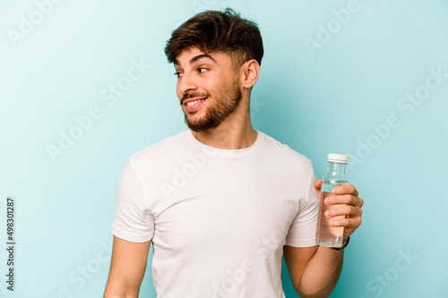 Young hispanic man holding a bottle of water isolated on white background looks aside smiling, cheerful and pleasant.