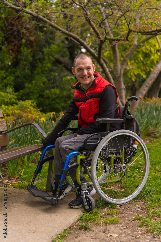 Portrait of a person with a disability in a red vest in a public park in the city. Sitting in the wheelchair next to a park chair Portrait of a person with a disability in a red vest in a public park in the city. Sitting in the wheelchair next to a park chair