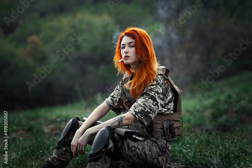 Sniper girl in a green field. Military snipers. The sniper smokes. Red-haired girl in camouflage. Fighting 