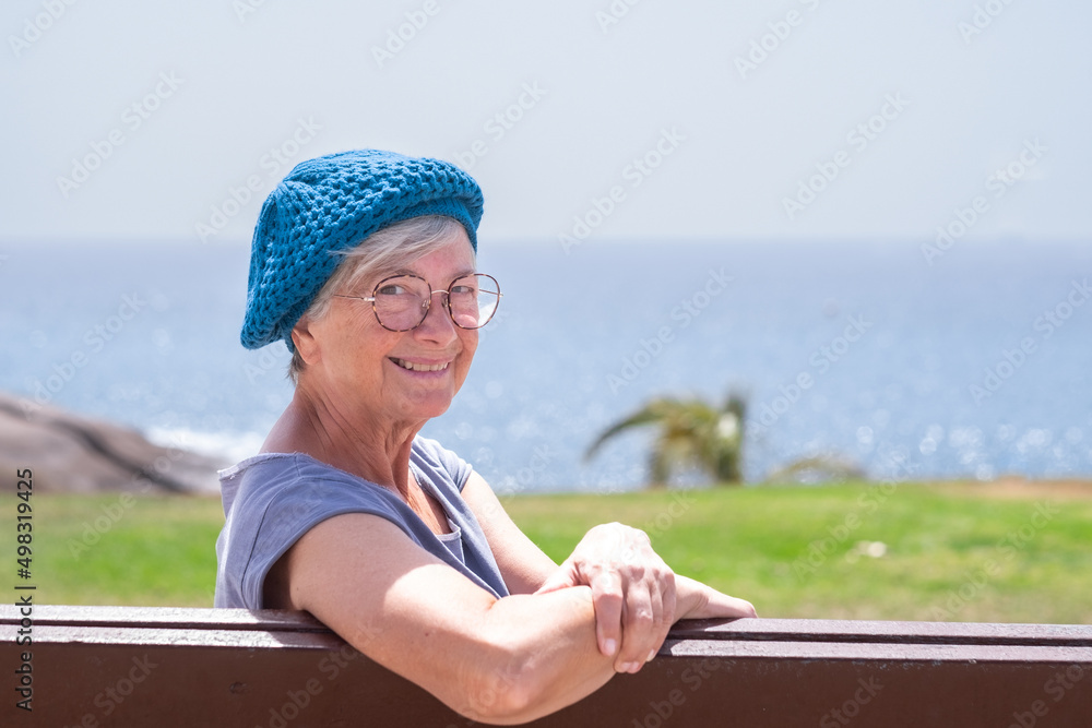 Smiling senior woman with blue cap sitting on bench close to the sea looking at camera. Elderly lady relaxing enjoying vacation or retirement under the sun. Horizon over water