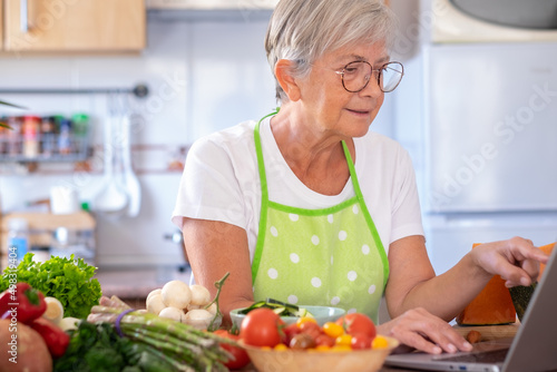Attractive senior woman in home kitchen with vegetables on the table looking for new menu on laptop. Caucasian modern elderly woman enjoying healthy eating