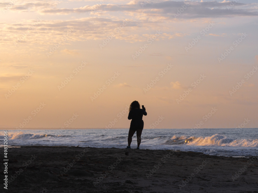 silhouette of a woman walking on the beach at sunset and taking a photo