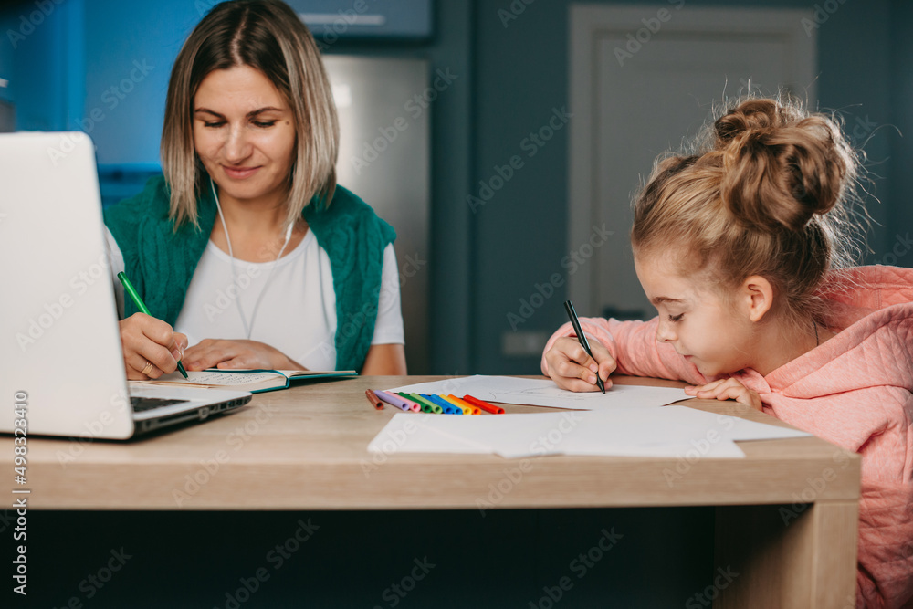 Woman with earphones working remotely on laptop while her daughter drawing with colourful pencils next to her. Freelance, online work, studying, learning with Woman with earphones working remotely on laptop while her daughter drawing with colourful pencils next to her. Freelance, online work, studying, learning with
