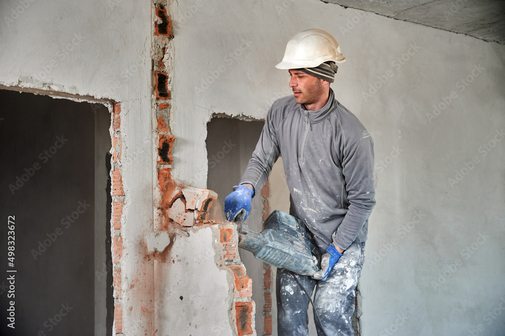 Worker in protective helmet with perforator completing deconstruction of rest of brick wall inside room. Builder carring out repair work on dismantling of walls and partitions. Worker in protective helmet with perforator completing deconstruction of rest of brick wall inside room. Builder carring out repair work on dismantling of walls and partitions.