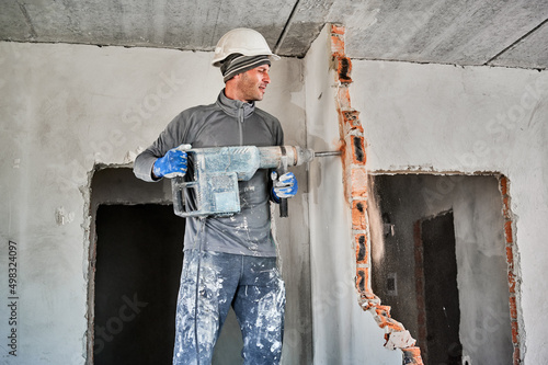 Side view of man builder in workwear drilling wall with hammer drill. Male worker using drill breaker while destroying wall in apartment under renovation. Demolition work and home renovation concept. photo