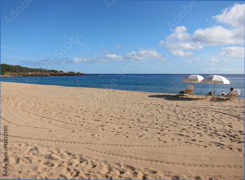 Sand full of footsteps and tire tracks. Deep blue sea, blue sky with white clouds. View of the coast. White beach umbrellas and chairs. Lanai, Hawaii, Hulopoe beach.