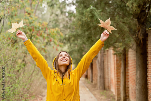 Front view of happy woman with raincoat raising arms with maple tree leaves. Horizontal mid waist view of woman looking up at fallen leaves in yellow hoodie outdoors. People and nature backgrounds.